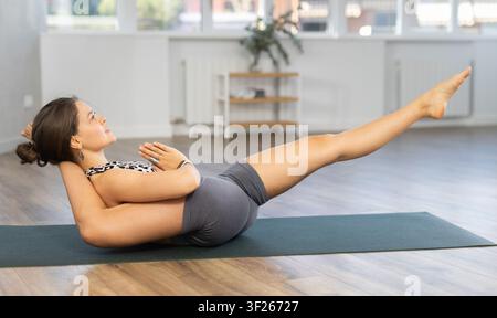 Young focused woman doing plough pose, variety of Halasana pose, difficult yoga pose in yoga studio Stock Photo