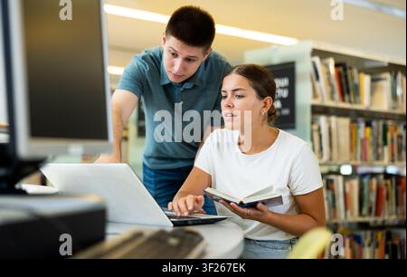 Young interested couple of students spending time together in library, reading books and browsing educational websites on laptop Stock Photo
