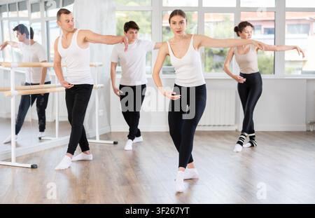 Experienced young female ballet dancer, dressed in white top and black leggings, executing dance moves with focused precision among group of people Stock Photo