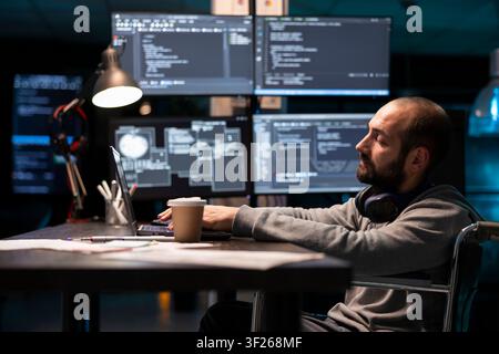 Male developer in wheelchair types on laptop while surrounded by large coding monitors in dim tech office. The scene highlights focus, software development, and accessible work environments. Stock Photo