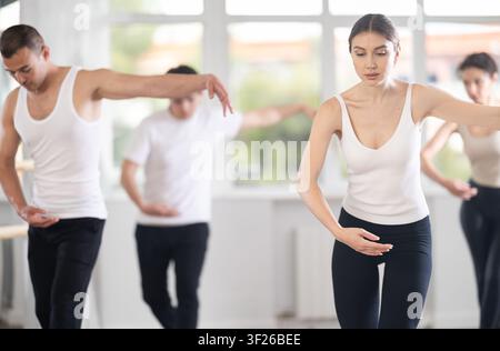 Experienced young female ballet dancer, dressed in white top and black leggings, executing dance moves with focused precision among group of people Stock Photo