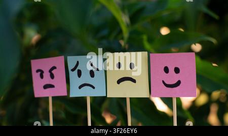 Four colorful sticky notes with different facial emotions placed in front of green leaves, suitable for mood, mental health, psychology, emotional exp Stock Photo