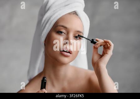 Young Woman Applying Mascara, Towel Wrapped Hair, Focused Expression, Home Bathroom, Beauty Concept Stock Photo
