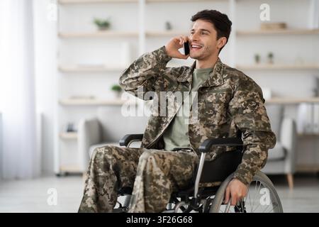Smiling Young Adult Male In Military Uniform Using Smartphone While In Wheelchair, Healthcare Concept Stock Photo