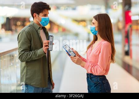 Young Man And Woman Using Smartphones In Mall, Wearing Face Masks, Shopping Concept Stock Photo