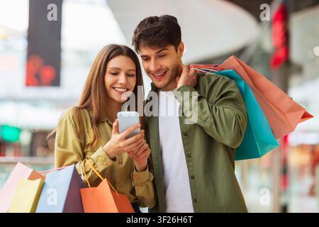 Happy Young Couple Shopping Together In Mall, Using Smartphone To Check Offers Stock Photo