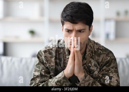 Worried Young Hispanic Man In Military Uniform Sitting On Couch, Indoor Setting, Mental Health Concept Stock Photo