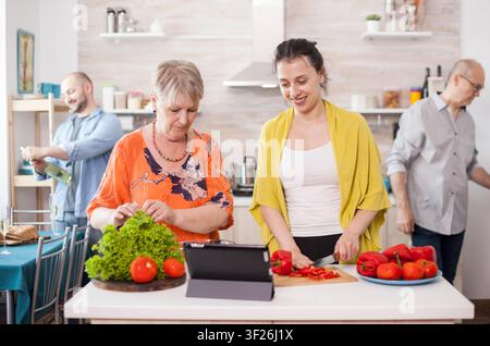 Mother and daughter follow a tablet recipe for healthy salad while family members prepare food together in a home kitchen in a realistic family lifestyle situation shaped by family and kitchen. Stock Photo