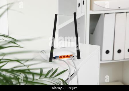 White wireless router with two antennas and connected ethernet cables on a desk in a modern office setting, the background features blurred white offi Stock Photo