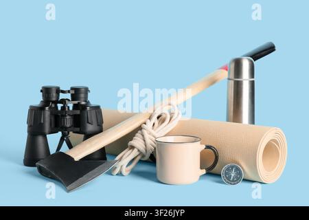 Camping and hiking gear with binoculars, axe, rope, mug, thermos, mat, and compass on a blue background Stock Photo