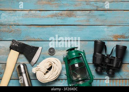 Camping and hiking gear with axe, compass, rope, thermos, lantern, and binoculars on a blue wooden background Stock Photo