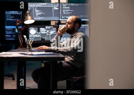Tired male employee drinks hot beverage while typing on laptop in security operations center. Focused programmer pushes through late night workload, surrounded by illuminated monitors and office tech. Stock Photo