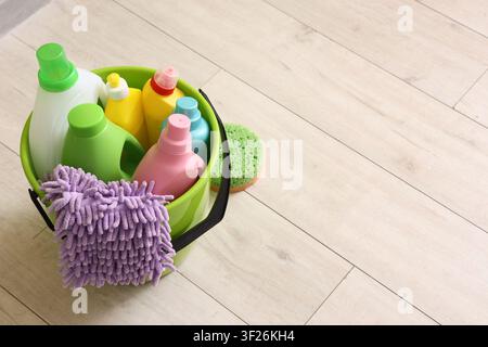 Green bucket filled with colorful cleaning supplies and a purple microfiber cloth on a light wooden floor, the items are arranged on a light wooden fl Stock Photo