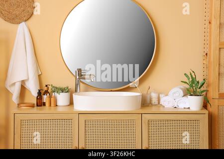 Modern bathroom interior with round mirror, white sink, wooden vanity, and personal care items on a peach wall Stock Photo