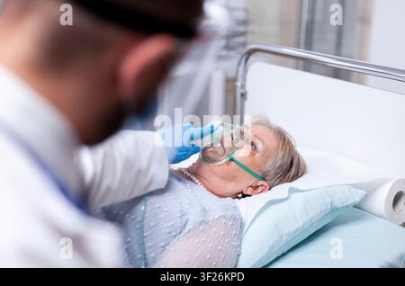 Doctor adjusts oxygen mask for senior hospital patient as safety precaution, oxygen mask and protective eyewear define the visible context in a hospital or clinic setting Stock Photo