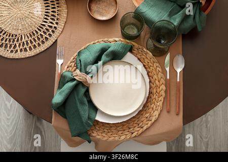 Elegant table setting with a woven placemat, ceramic plates, green napkin, and modern cutlery on a brown tablecloth Stock Photo