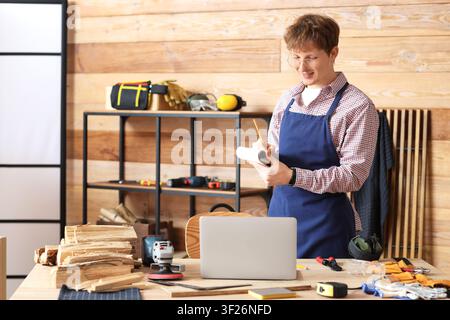 Young carpenter with notebook using laptop at table in workshop Stock Photo