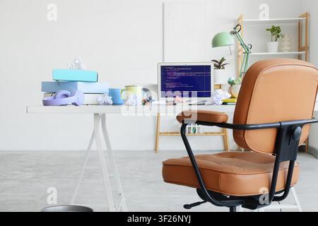 Modern home office workspace with laptop displaying code, messy desk, and brown leather chair, remote work Stock Photo