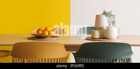 Modern dining room interior with yellow and white walls, wooden table, colorful chairs, fruit bowl, and tea set Stock Photo
