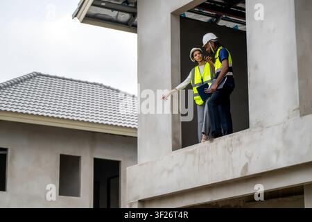 Construction managers holding clipboard and tablet pointing down from second floor balcony of unfinished residential house, concept of teamwork and pr Stock Photo