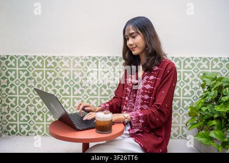 Asian woman using a laptop while sitting at a cafe table with iced coffee. Remote work, freelance and digital nomad lifestyle concept. Stock Photo