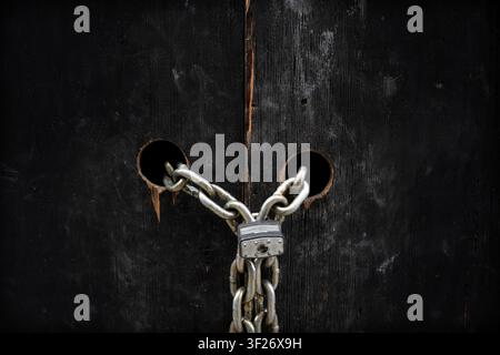 Close-up of a heavy metal chain and padlock securing weathered black wooden doors. The image conveys security, protection, restricted access, privacy, safety, and resilience with a dramatic rustic texture. Stock Photo