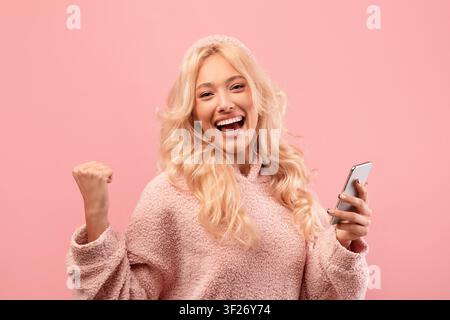 Happy Young Woman Celebrating With Smartphone In Hand, Joyful Expression, Pink Background Stock Photo