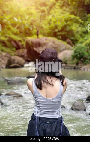 Back view of a young asian woman wearing a bucket hat and tank top, standing by a river in a lush green forest Stock Photo