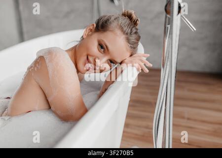 Young Woman Relaxing In Bathtub With Bubbles, Smiling At Home, Wellness Concept Stock Photo