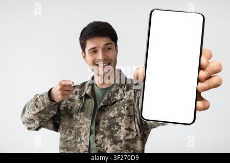 Smiling Young Man In Military Uniform Holding Smartphone With Empty Screen, Military Concept Stock Photo