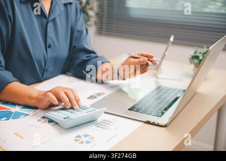 Business analyst using calculator and laptop with printed financial charts on a bright office desk, showing accounting, budget planning, data analysis Stock Photo