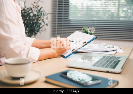 Business analyst writing on financial report while working with laptop, smartphone, coffee, and charts on office desk, representing accounting, market Stock Photo