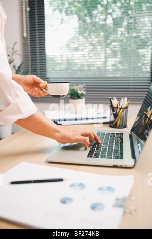 Professional working on laptop while holding coffee in modern office, with charts and documents on desk, representing business productivity, financial Stock Photo