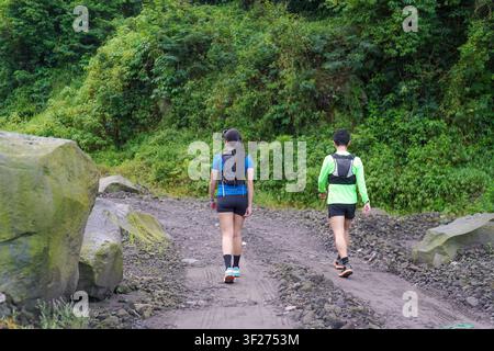 Two hikers walking side by side on a rocky trail surrounded by lush forest, wearing sporty outdoor gear. Stock Photo