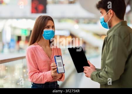 Woman Holding Smartphone With Qr Code And Man Holding Tablet In Mall, Masked Interaction Stock Photo