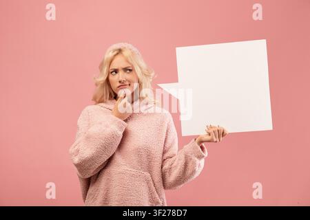 Thoughtful Young Woman Looking Up While Holding Blank Speech Bubble, Pink Background Stock Photo