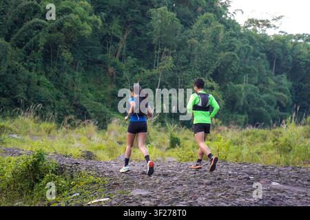 Asian Indonesian couple seen from behind as they jog together on a rocky trail in a lush green forest, living an active and sustainable lifestyle. Stock Photo