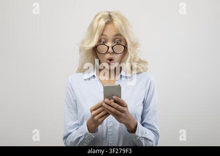Surprised Young Caucasian Woman With Glasses Holding Smartphone, Office Setting Stock Photo