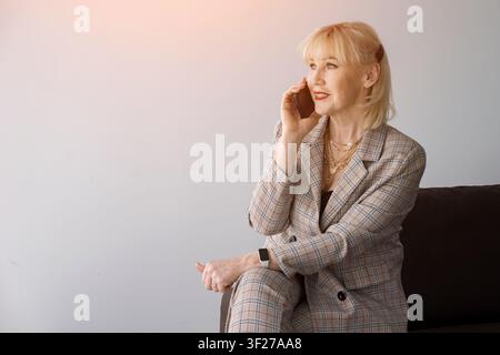 Senior blonde businesswoman seated indoors, smiling confidently while on a professional smartphone call, wearing a stylish plaid suit and engaged in Stock Photo
