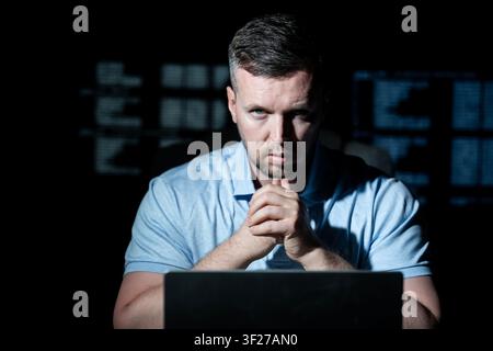 Hacker works on laptop at night. Hacker analyzes code screen in dark room. Hacker monitors network for cyber attack. Hacker targets data system with Stock Photo