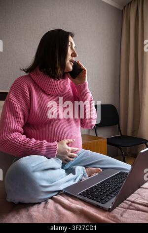 Pregnant woman sitting on bed at home, resting her hand on her face while using a laptop Stock Photo