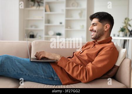 Smiling Young Caucasian Man Working On Laptop While Relaxing At Home Sofa Stock Photo