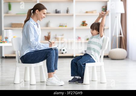 Child Raising Hands And Smiling, Female Therapist Taking Notes In Bright Office Setting Stock Photo