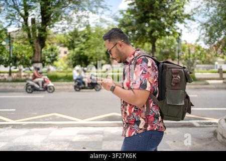Side view of an Indonesian man walking with a smartphone and backpack, showcasing mobile technology, commuting, and modern urban lifestyle. Stock Photo