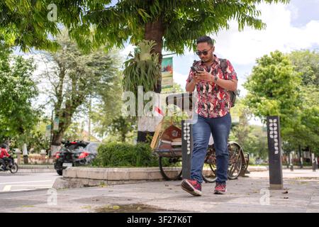 Asian Indonesian man with backpack using smartphone while walking on a city sidewalk surrounded by trees. Mobile technology, communication and urban l Stock Photo