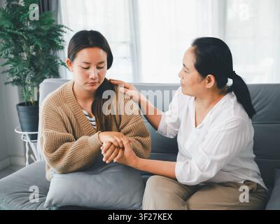 Middle aged Asian woman comforting sad adult daughter by holding hands on sofa at home, concept of emotional support, family care, empathy, counseling Stock Photo