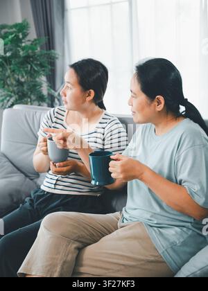 Asian mother and adult daughter sitting on sofa with cups at home, having conversation and pointing outside, concept of family communication, support, Stock Photo