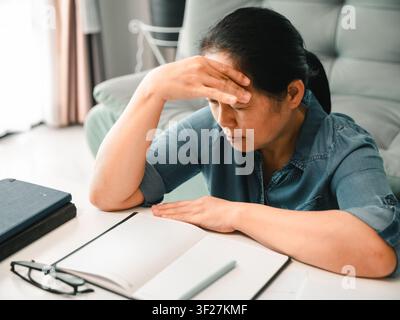 Asian woman sitting at table with notebook and glasses, touching forehead from headache, concept of stress, study, paperwork, work pressure, eye strai Stock Photo