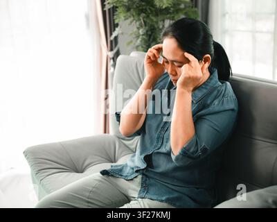 Middle aged Asian woman touching temples with eyes closed on sofa at home, concept of migraine, headache, stress, anxiety, burnout, fatigue, and healt Stock Photo