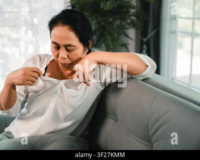 Asian woman feeling overheated and uncomfortable while sitting on sofa at home, pulling white shirt collar, concept of menopause, hot flash, sweating, Stock Photo